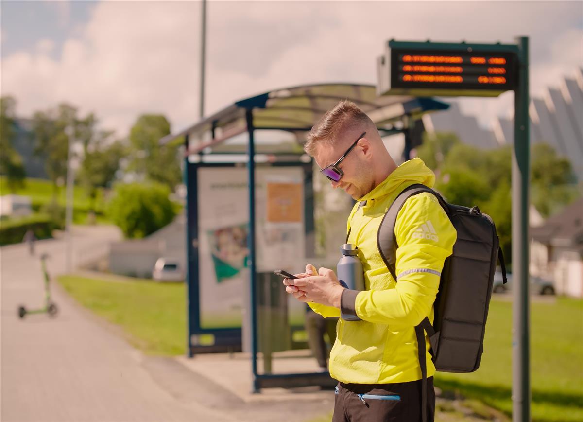 Mann i treningsklær foran busstopp. Sanntidsinformasjonsskjerm i bakgrunnen. - Klikk for stort bilde